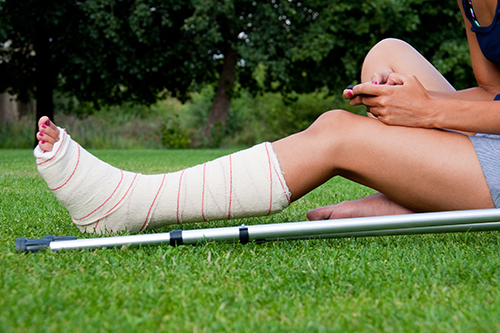 Leg in plaster of a girl sitting on the grass writing a text message with her smartphone. Crutches lying down at her side.