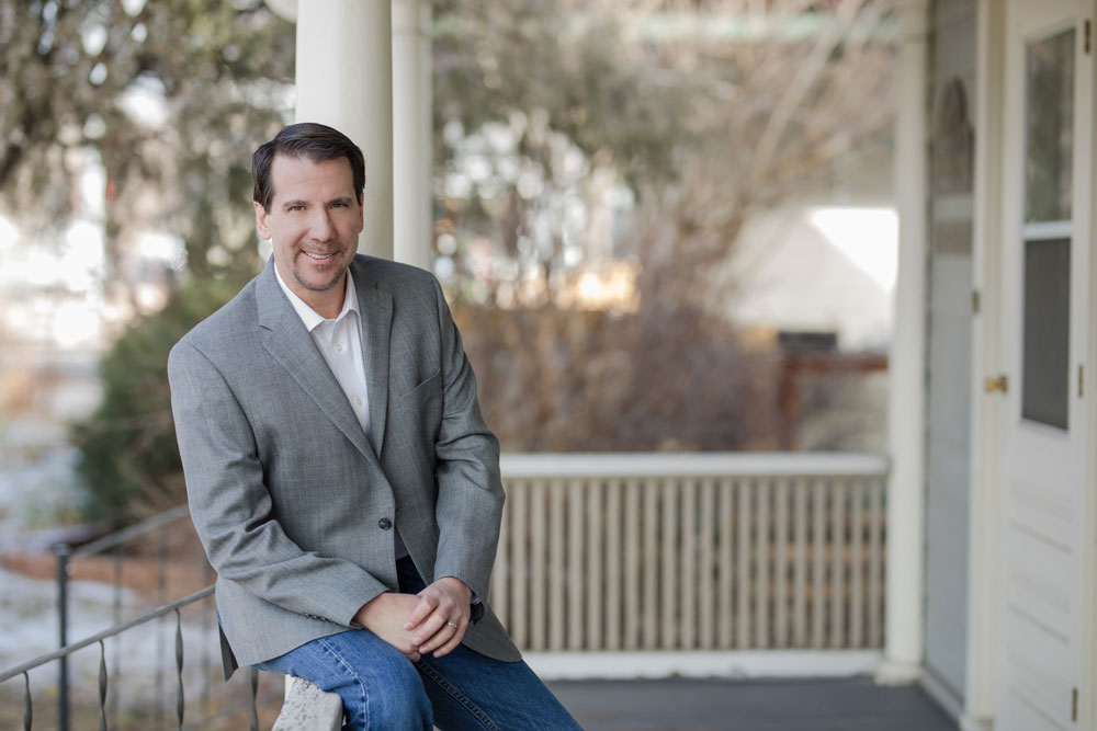 Greg Green of the Green Law Firm, P.C. sitting on a white porch while wearing a gray suit coat